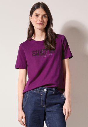 Paarse katoenen t-shirt met korte mouwen, voorzien van een zwart grafisch ontwerp en de tekst "VIND JE INNERLIJKE VREDE." Gecombineerd met donkerblauwe jeans.