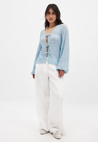 Hellblauer gehäkelter Cardigan mit langen Ärmeln und V-Ausschnitt, der einen Bindegürtel in der Taille hat, getragen über weißen, weit geschnittenen Leinenhosen.