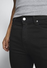 Persona vistiendo jeans negros de Calvin Klein y una camiseta sin mangas gris, con la mano relajada a su lado frente a un fondo liso.