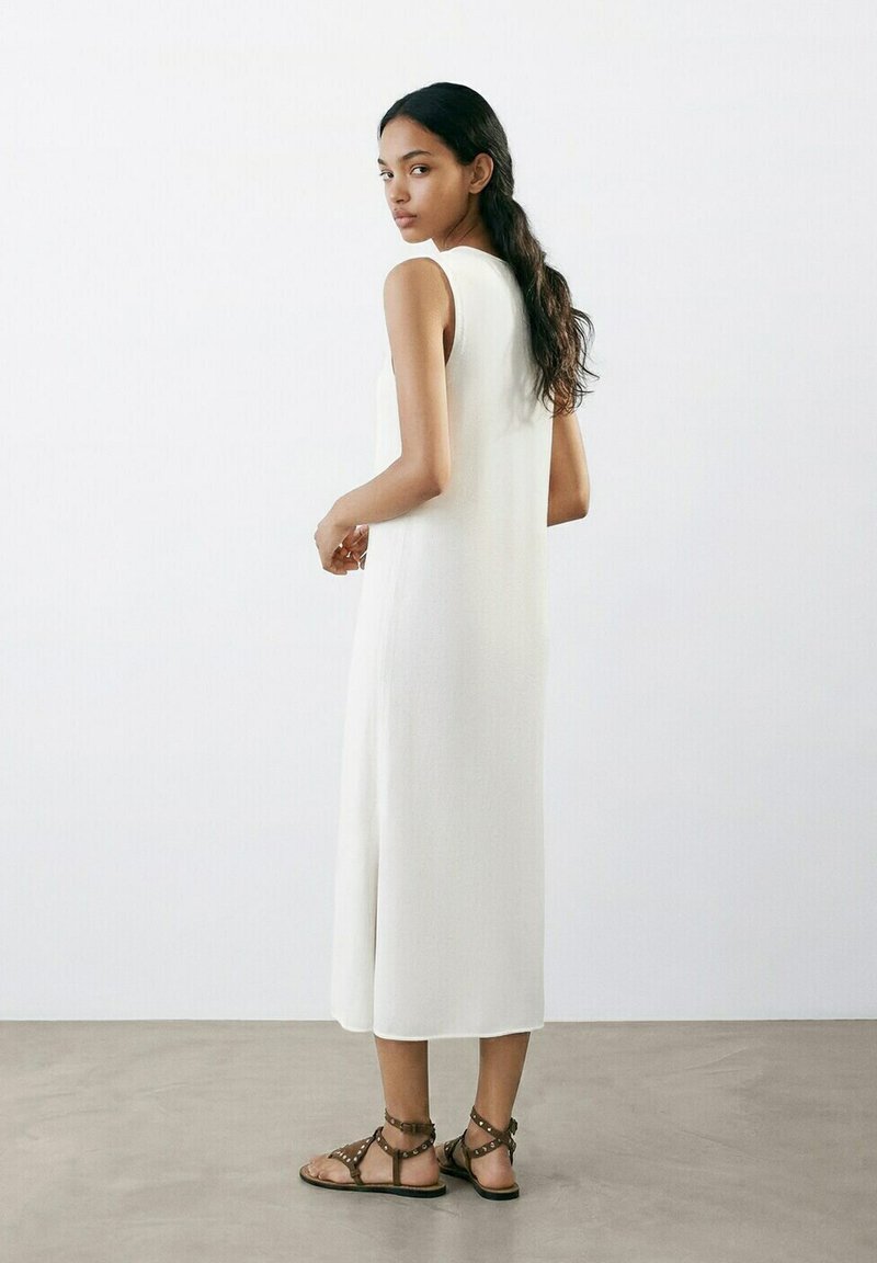 Donna con capelli lunghi e scuri che indossa un vestito midi bianco senza maniche e sandali bassi marroni con cinturini, in piedi su un pavimento grigio.