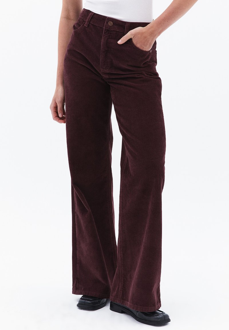 Un pantalon en velours côtelé de couleur bourgogne avec une coupe large, présentant des côtes verticales et des poches avant. Associé à des chaussures noires à bout rond.