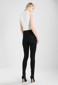 Jeans negros ajustados de cintura alta, con tejido suave y bolsillos traseros, combinados con una blusa ajustada de color claro y tacones altos.