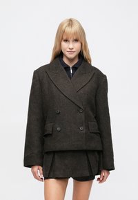 Sötétbarna, dupla soros kabát texturált anyagból, két első zsebbel és hangsúlyos hajtókával, fekete felső és szoknya fölött viselve.
