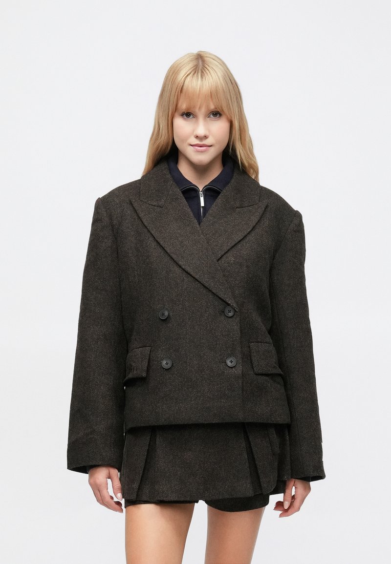 Sötétbarna, dupla soros kabát texturált anyagból, két első zsebbel és hangsúlyos hajtókával, fekete felső és szoknya fölött viselve.