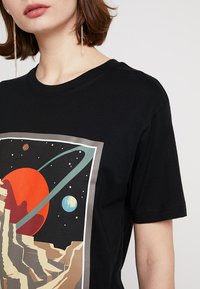 Sort bomulds T-shirt med et livligt rummet grafik, der viser en rød planet, ringe og to mindre planeter, indrammet af en rektangulær kontur.