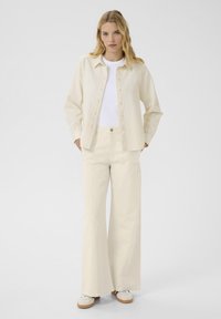 Beige linnen overhemdjack met knopen, gecombineerd met bijpassende wijde broek. Witte t-shirt eronder, gestyled met witte sneakers.