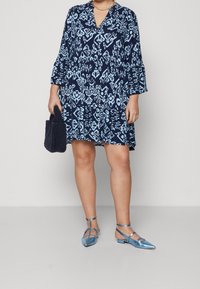 Kvinna i en marinblå mönstrad klänning, med en mörk texturerad handväska och metallicblå sandaler med låg klack.