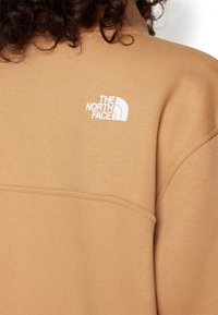 Personne aux cheveux bouclés portant un sweat North Face beige, logo blanc visible en haut du dos près de l'épaule gauche.