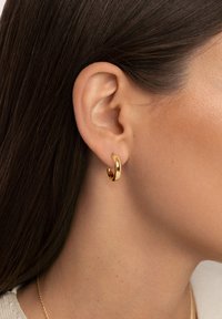 Pendiente de aro dorado con un acabado liso y pulido. Diseño curvado y tamaño mediano, que destaca por su textura brillante y una sutil calidad reflectante.