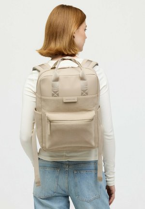 Vrouw met kort haar die een beige ribfluwelen rugzak draagt over een wit shirt met lange mouwen en lichtblauwe spijkerbroek, met haar rug naar de camera toe.