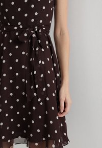 Robe marron à pois avec une taille cintrée, un détail nœud papillon et une jupe évasée. Confectionnée en tissu léger ; présente des motifs circulaires blancs.