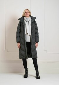 Longue doudoune gris foncé avec un col montant et une fermeture à boutons sur le devant. Associée à un pull gris clair, un pantalon noir et des bottines noires.