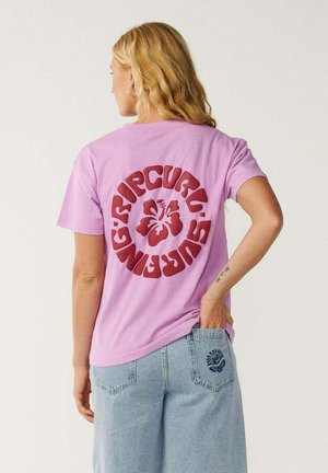 Vrouw met blond haar die een roze t-shirt draagt met rode “Rip Curl Surfing” tekst en bloemenlogo, lichtblauwe spijkerbroek met ontwerp op de achterzak.