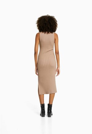 Femme aux cheveux bouclés portant une robe midi sans manches côtelée beige et des bottines noires, debout de dos sur un fond blanc.