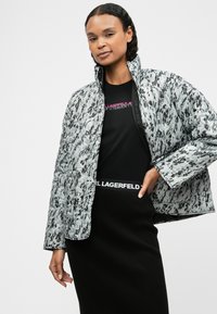 Rīvēta pūkainā jaka pelēkā krāsā ar melnu abstraktu rakstu, ar augstu apkakli, ar rāvējslēdzēju priekšā un divām sānu kabatām.