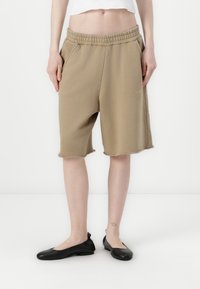 Beżowe szorty do kolan z elastycznym paskiem w talii i kieszeniami po bokach. Surowe krawędzie u dołu i minimalne detale logo na nogawce.