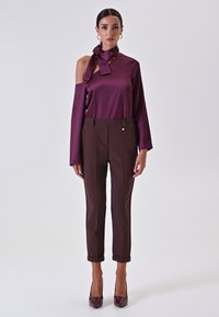 Blusa di satin bordeaux con dettaglio al collo annodato, abbinata a pantaloni marroni sartoriali, con orli a risvolto e un accento lucido sulla vita.
