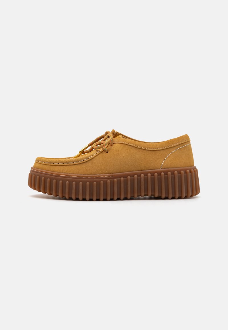 Chaussures en daim marron avec un bout arrondi, des coutures complexes et une semelle en caoutchouc épaisse et striée de couleur marron foncé. Elles possèdent des lacets.