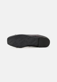 Semelle de chaussure noire en caoutchouc texturé, présentant un nom de marque embossé et des motifs. Talon légèrement surélevé, bords lisses et design minimaliste.