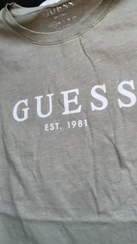 T-shirt de algodão cinza com o logótipo branco "GUESS" e texto "EST. 1981". Design de gola redonda com um tecido suave, texturizado e com mínimas rugas.