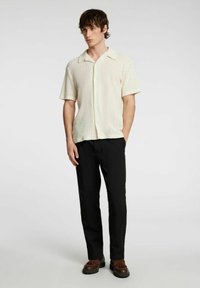 Kremowa koszula krótko rękawowa z guzikami, o teksturowanym wzorze, zestawiona z czarnymi spodniami i brązowymi butami. Prosty krój kołnierzyka, luźny fason.