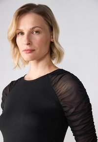 Top negru cu un corp ajustat și mâneci lungi transparente, pliate. Textură netedă cu detalii moi de drapaj la umeri.