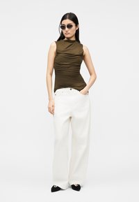 Nő barna ujjatlan fodros felsőt, fehér bőszárú nadrágot, fekete hegyes cipőt és kis fekete napszemüveget visel, egyik kezét zsebre dugva áll.