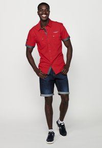 Camisa de color rojo con detalles grises, dos bolsillos en el pecho y mangas enrolladas, combinada con pantalones cortos de mezclilla y zapatillas de deporte azul marino.
