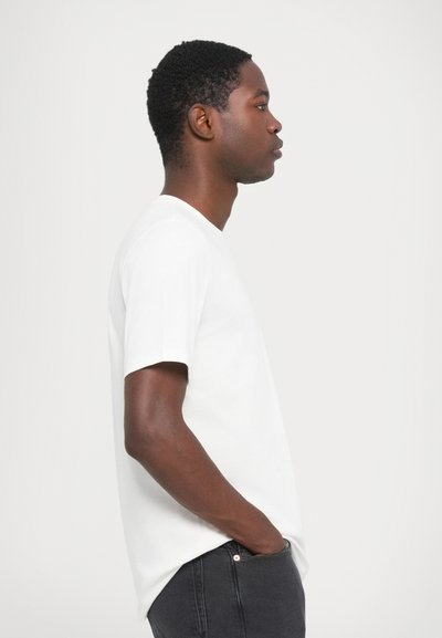 T-shirt bianca in cotone con maniche corte, collo rotondo e vestibilità rilassata. Abbinata a jeans grigio scuro, mostrata di lato.