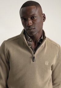 Sudadera con cremallera en color caqui, hecha de un tejido de canalé texturizado, con un pequeño parche de logo y una camisa de color gris debajo.
