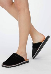 Schwarze Slippers zum Hineinschlüpfen mit weichem Wildleder-Obermaterial, flacher Gummisohle und kontrastierendem weißen Nähten entlang der Kanten. Strukturierte Oberfläche.