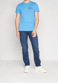 Muž v světle modrém tričku Tommy Hilfiger, tmavě modrých džínách a bílých teniskách, stojící na jednobarevném bílém pozadí.