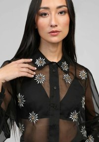 Blouse noire transparente avec col, boutons sur le devant et broderie florale avec des accents en argent et or sur le tissu.