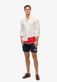 Camisa de lino blanca ligera, pantalones cortos de baño rojos y azules marino con logo, zapatos de barco marrones. El modelo se presenta de manera confiada con las manos en los bolsillos.