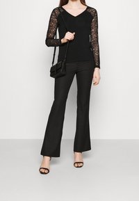 Zwarte top met lange mouwen en kantmouwen, met een V-hals, gecombineerd met zwarte flarebroeken en zwarte sandalen met bandjes. Kleine crossbody tas.