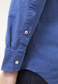 Camicia blu con bottoni e tessuto strutturato. Presenta bottoni bianchi con dettagli rossi ai polsini e all'orlo. Design slim fit, con dettagli cuciti.