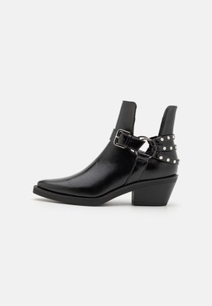 Bottine en cuir noir avec bout pointu, talon bloc, bride à boucle et lanières décoratives cloutées à l'arrière.