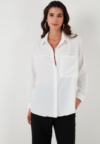 White button-up shirt made of lightweight fabric with a single chest pocket, long sleeves, and classic collar. Smooth texture, relaxed fit.