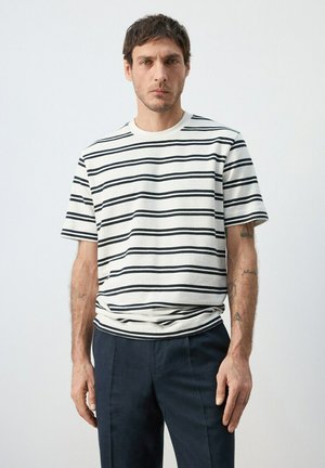 Hombre con camisa blanca de manga corta con rayas horizontales negras y pantalones oscuros, de pie frente a un fondo claro y uniforme.