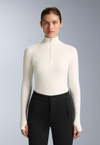 Kvinde iført en tætsiddende hvid kabelstrikket sweater med en halvlange lynlåsskrave og sorte højtaljede bukser med lynlåslommer, stående mod en grå baggrund.