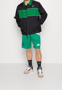 Zwarte en groene windbreaker met een logo, groene shorts met witte en paarse accenten, zwarte sokken en sneakers met grijze en zwarte details.