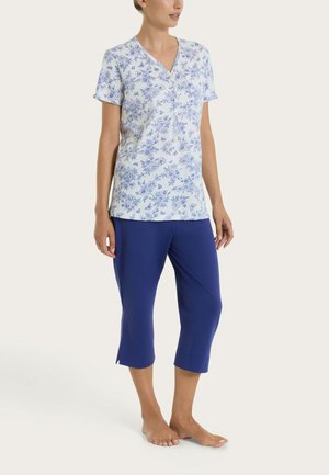 Donna che indossa una maglietta bianca a maniche corte con stampa floreale blu e pantaloni corti blu tinta unita, in piedi a piedi nudi su uno sfondo chiaro.