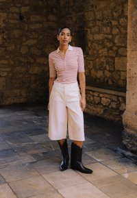 Femme debout contre un mur en pierre, portant un polo rayé, des culottes blanches larges et des bottes noires mi-mollet sur un sol carrelé.