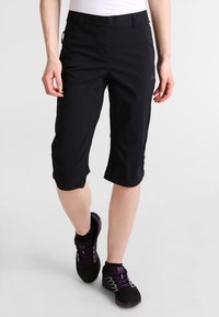 Kvinna som bär svarta knälånga shorts och svarta sportskor med lila snören, som går mot en enkel vit bakgrund.