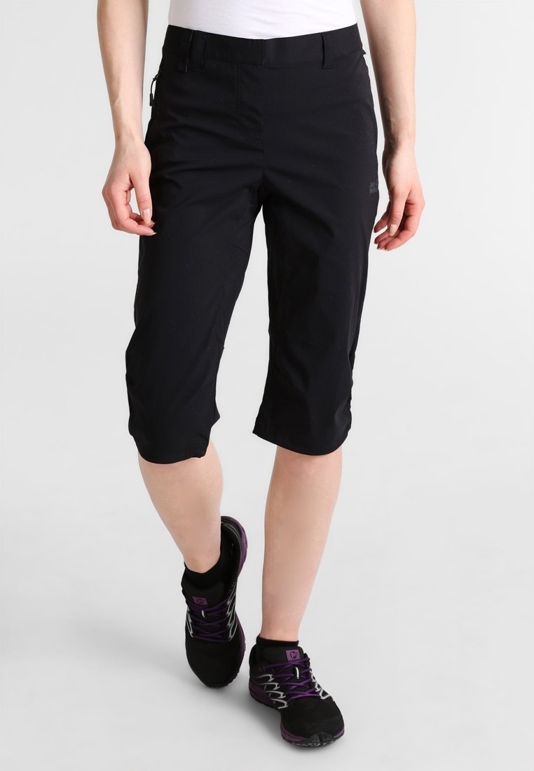 Kvinna som bär svarta knälånga shorts och svarta sportskor med lila snören, som går mot en enkel vit bakgrund.