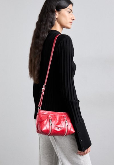 Rote Leder-Umhängetasche mit silbernen Reißverschlüssen und Akzenten, in strukturierter Form und mit verstellbarem Tragegurt, gehalten von einem Model.