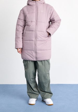 Persoon die een lichtpaarse gewatteerde jas met capuchon draagt, groene cargobroek en witte sneakers, staand met één hand in de zak.