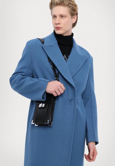 Modrá vlnená kabát s dvojradovým designom, veľkými klopami a hladkým povrchom; doplnený čiernou koženou taškou cez plece so striebornými doplnkami.