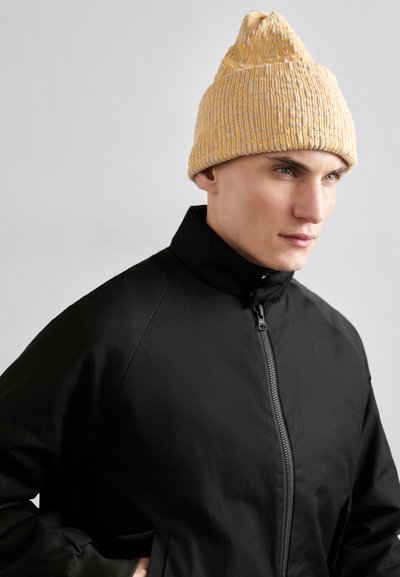 Gelbe Strickmütze mit strukturiertem Muster und spitzem oberen Ende, kombiniert mit einer schwarzen Zip-Jacke mit hohem Kragen.