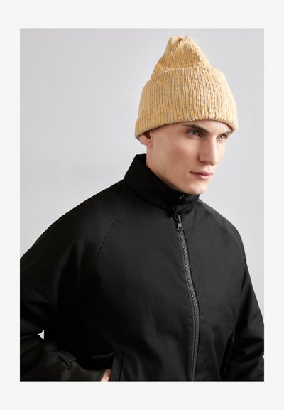 Gelbe Strickmütze mit strukturiertem Muster und spitzem oberen Ende, kombiniert mit einer schwarzen Zip-Jacke mit hohem Kragen.